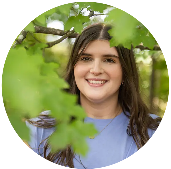 A female student wearing a lilac sleeveless top stands among green leafy tree branches.