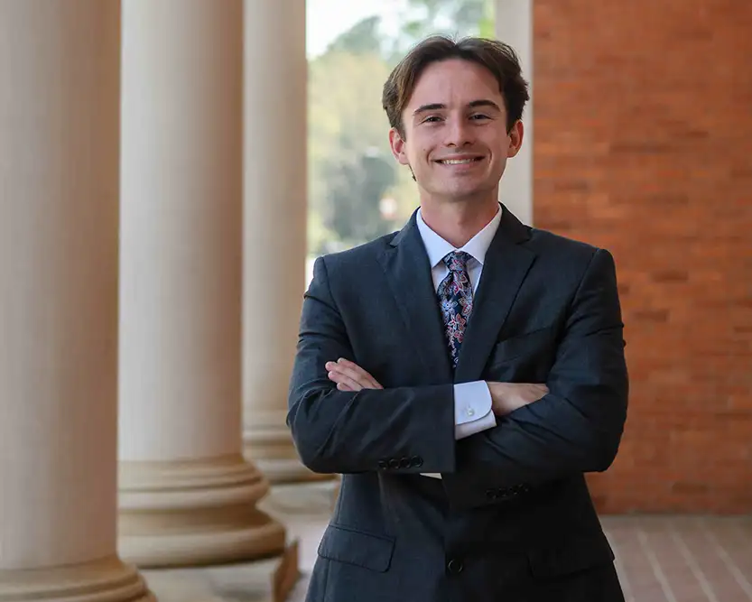 A smiling male college student in a dark suit and patterned tie stands with his arms crossed at a building entrance lined with large stone columns and a brick wall.