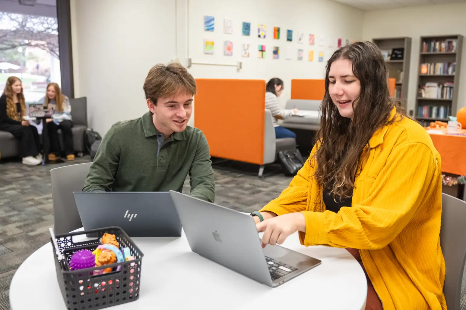 A male college student and a female college student sit at a table in a campus learning center, working together on laptops while other students study in the background.