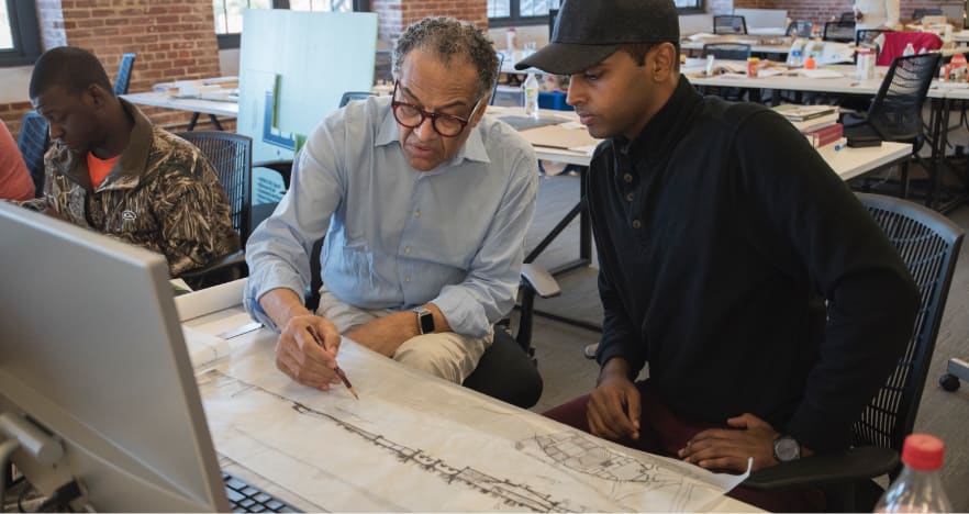Elderly male instructor and male college student inspect bridge design printed on long paper in an academic building.