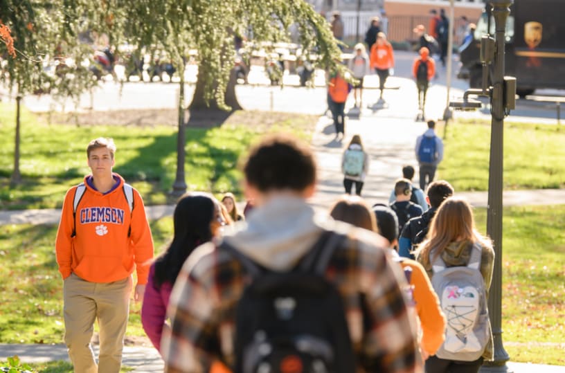 Students walk on a sidewalk across a green space separating academic buildings.