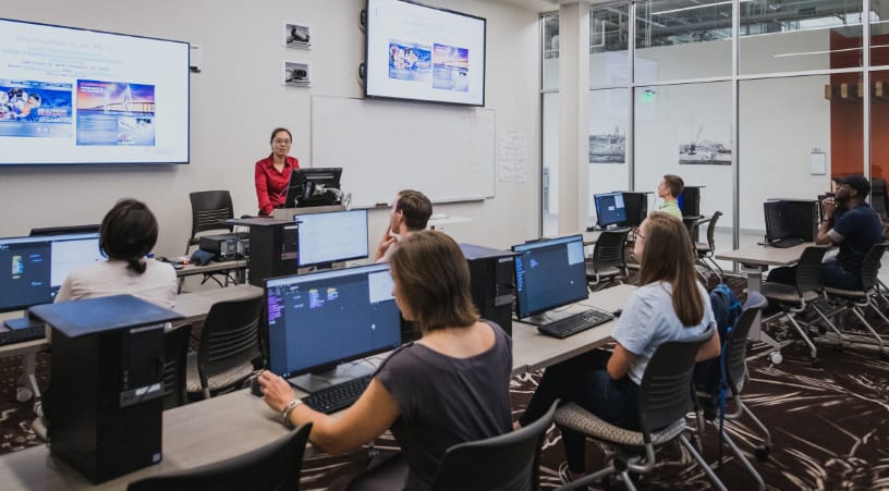 Two rows of students sit in at desktop computers while listening to their professor’s lecture.