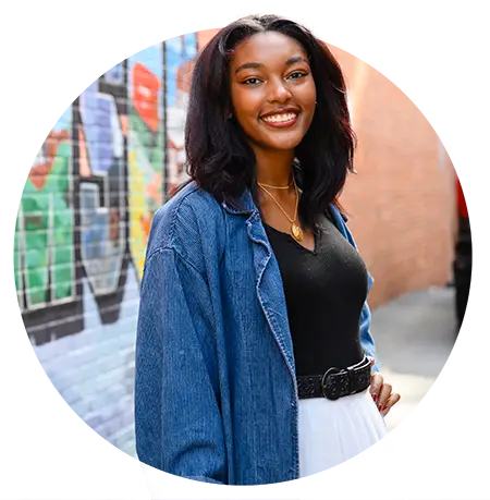 A female student wearing a white skirt, black top and navy corduroy jacket poses in front of a colorful Clemson postcard mural.