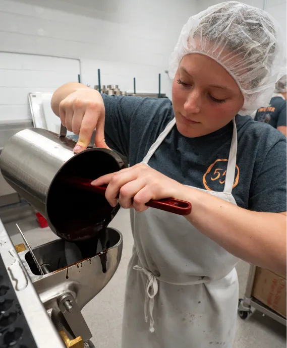 A female student wearing a T-shirt, apron and hairnet pours a liquid mixture into a machine.