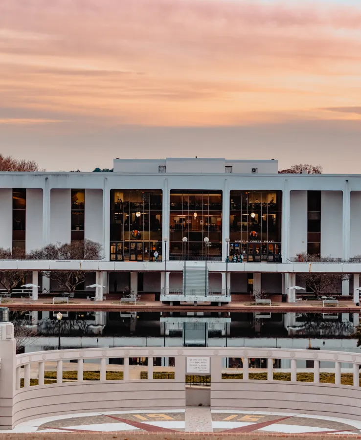 A white concrete college library building with large glass windows sits in front of a reflection pond and amphitheater at sunset.