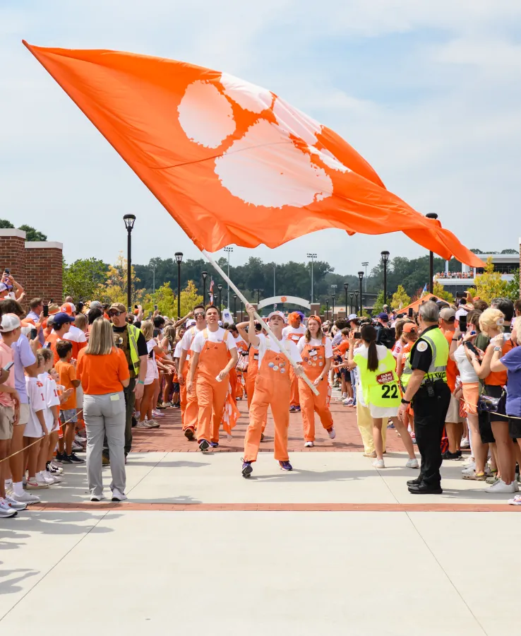 A group of college students wearing white T-shirts and Clemson Orange overalls lines up on a concrete and brick pathway lined with Clemson fans. One student waves a large orange flag adorned with a white Clemson Tiger Paw.