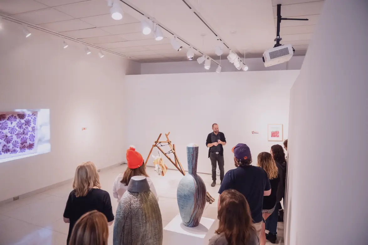 A man stands in a brightly lit white gallery space next to an abstract sculpture as a group of people looks on and stands among two ceramic works on display.