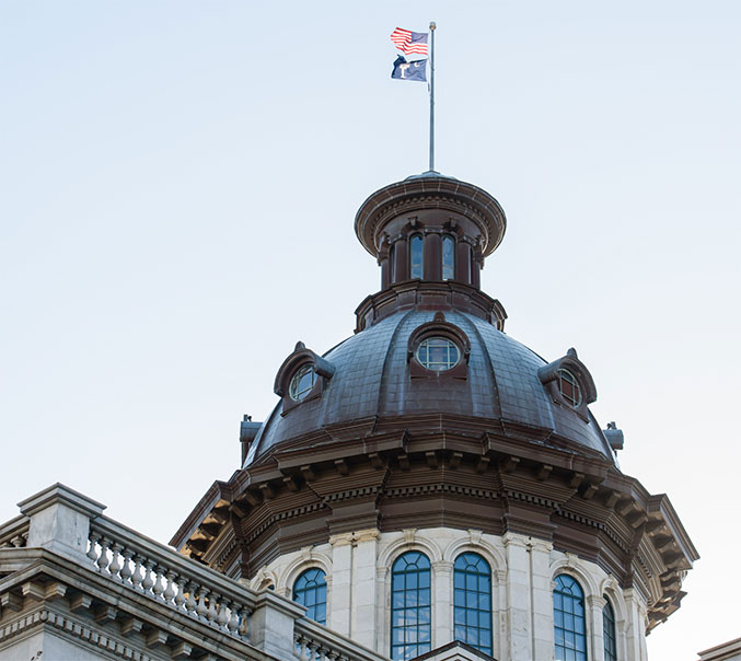 The US and state flag fly in the wind above South Carolina's capitol building.