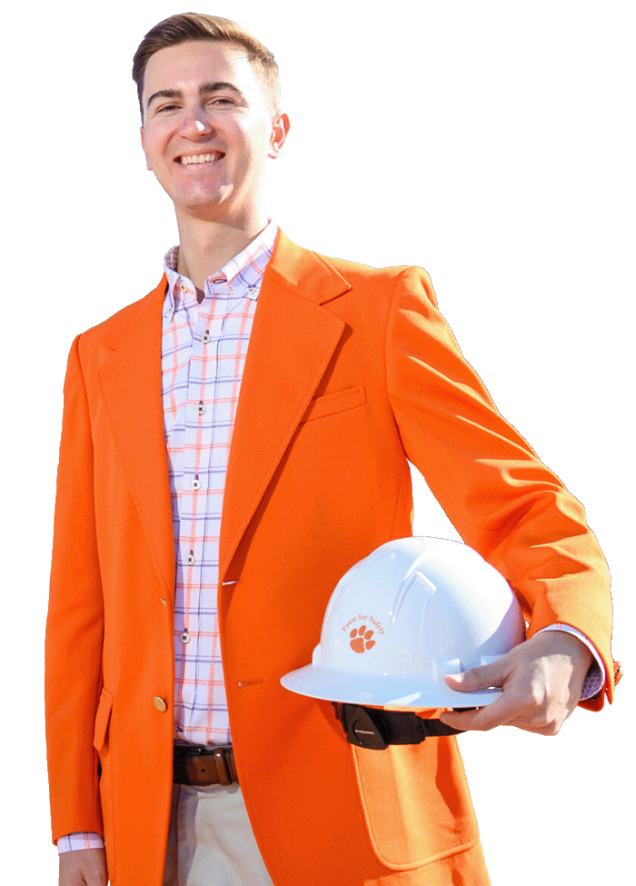A male college student wearing khaki pants, an orange-and-purple checkered button-down shirt and an orange blazer holds a white construction hard hat and stands in front of the Tiger Walk archway entrance constructed from brick and concrete.