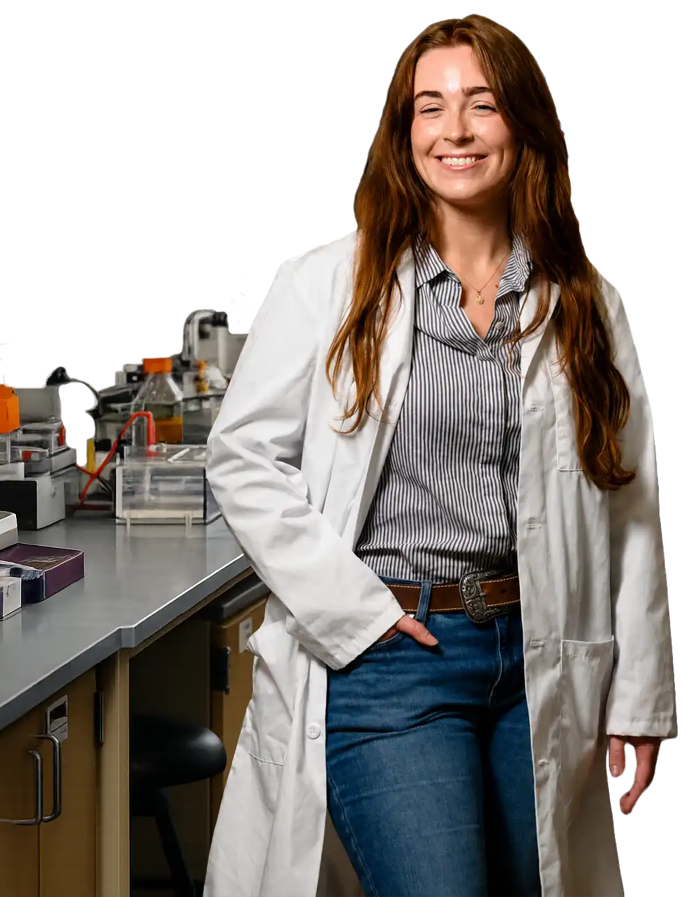 A female student wearing jeans, a button-down shirt with black-and-white stripes and a white lab coat stands and smiles in a laboratory between shelves and work stations of research equipment.