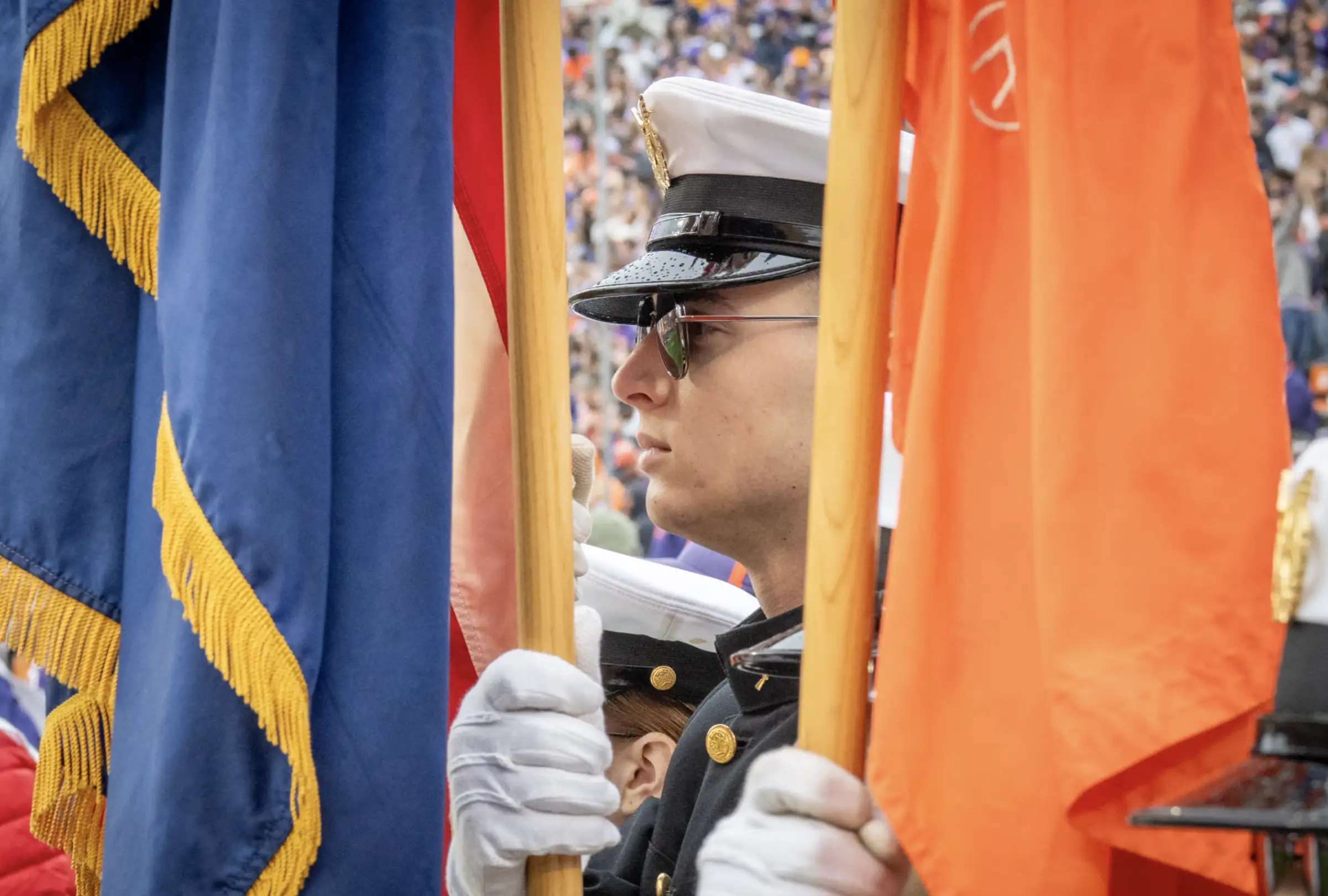 A Clemson student wearing a military uniform and sunglasses presents the flag with a large crowd in the background.