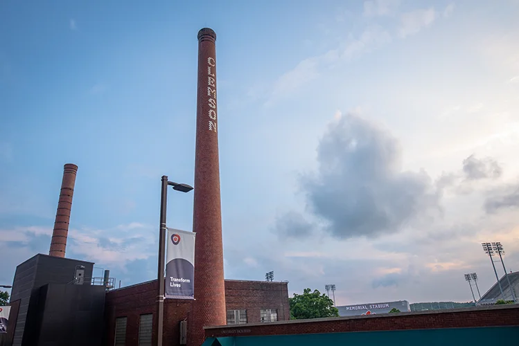 Two smoke stacks, with one that says, 'Clemson' in white vertical letters, rise in front of brick buildings with the lights and upper deck of a football stadium in the background.