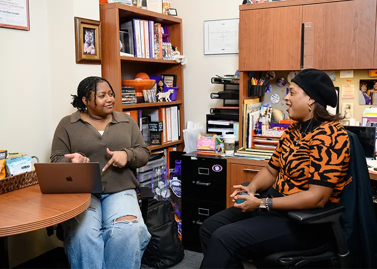 A female student wearing jeans and a brown long-sleeved sweater and a female staff member wearing black pants and a tiger-stripe short-sleeved sweater and a black hat sit together in an office space.