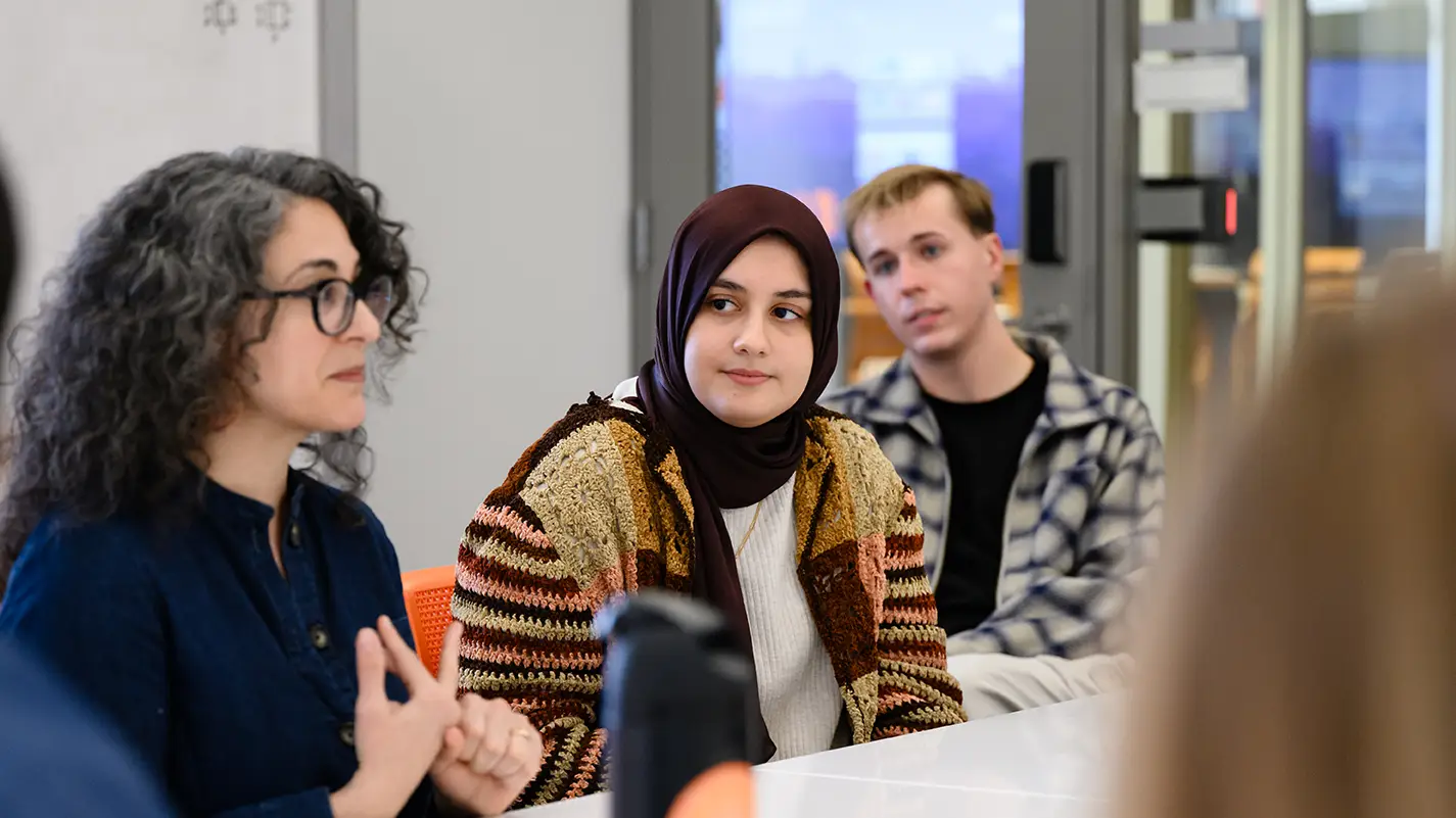 A small group of people sit around a conference table in a classroom meeting room. In the forefront, a woman wearing a navy sweater and glasses gestures while speaking. Next to her, a female student wearing a brown headscarf and multicolored patchwork cardigan listens, while a male student wearing a plaid shirt looks on from behind.