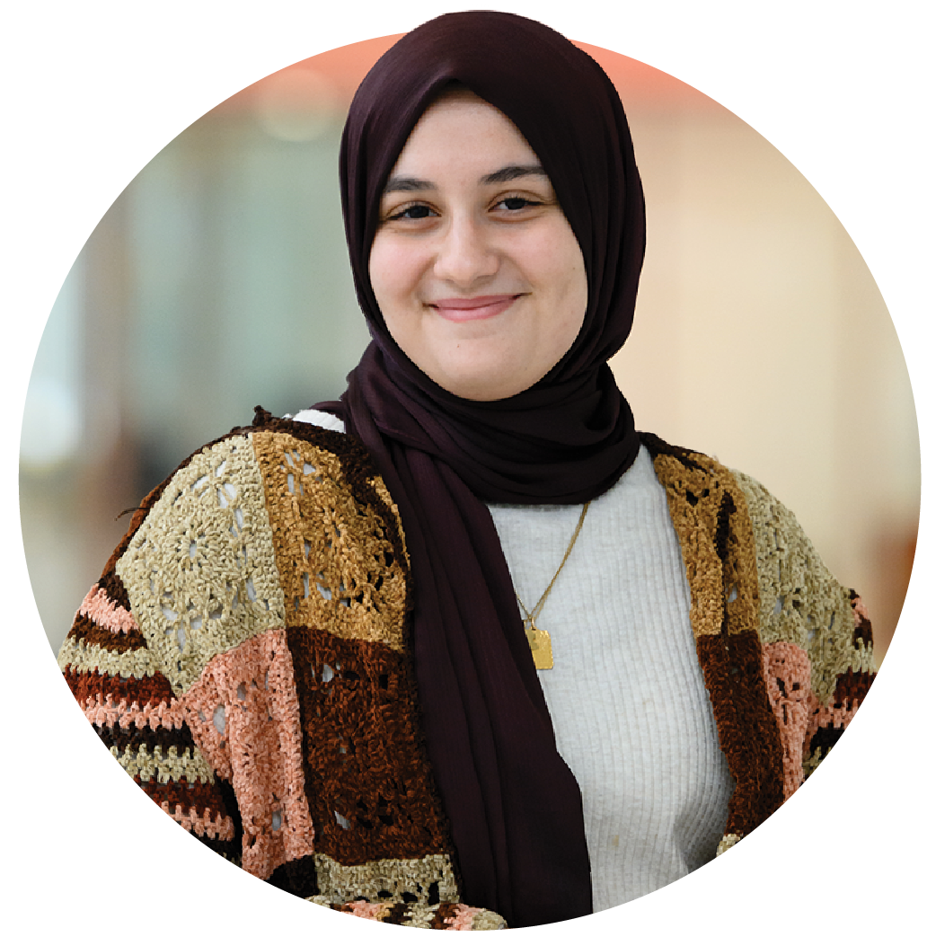 A female student wearing a burgundy headscarf, a multicolored patchwork cardigan sweater over a cream-colored shirt and brown pants poses in the atrium of an academic building.