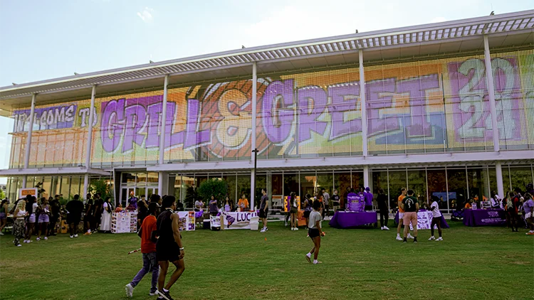 Groups of students at tables on a lawn in front of a building with large signage taking up the entire side of the building that reads, Grill & Greet in purple and orange.