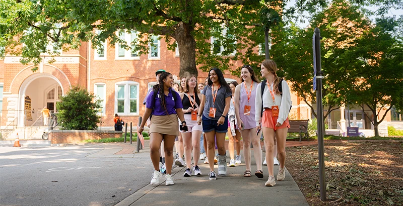 A female student wearing a khaki skirt and purple polo leads a group of new undergraduate students down a sidewalk in front of a brick academic building.