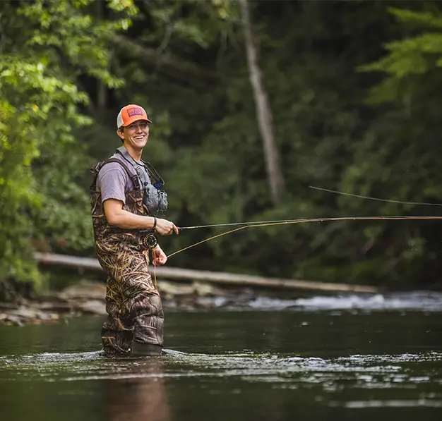 A male student wearing an orange baseball cap, light purple T-shirt and camouflage print fishing waders stands in a stream and holds a fishing pole.