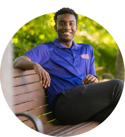 A male student wearing a purple polo, gray pants and black-and-white checkered shoes sits on a bench between two green leafy trees.