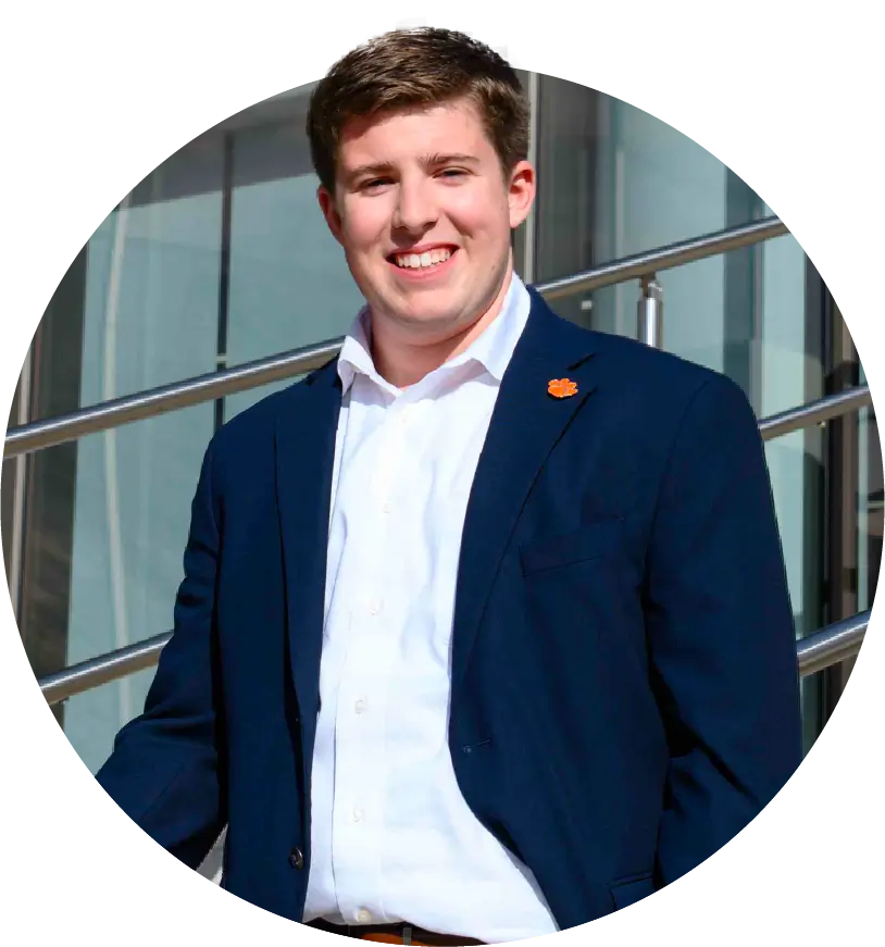 A male student wearing a white shirt and navy blazer poses outdoors on the steps of a campus building. 