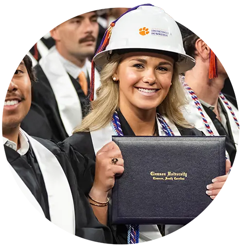 A female student wearing a white construction hard hat and a black graduation gown holds a Clemson University diploma.