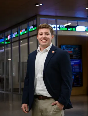 A male student wearing khaki pants, a white button-down shirt and a navy blazer with an orange Tiger Paw lapel pin stands in an academic building with a stock ticker illuminated in the background.