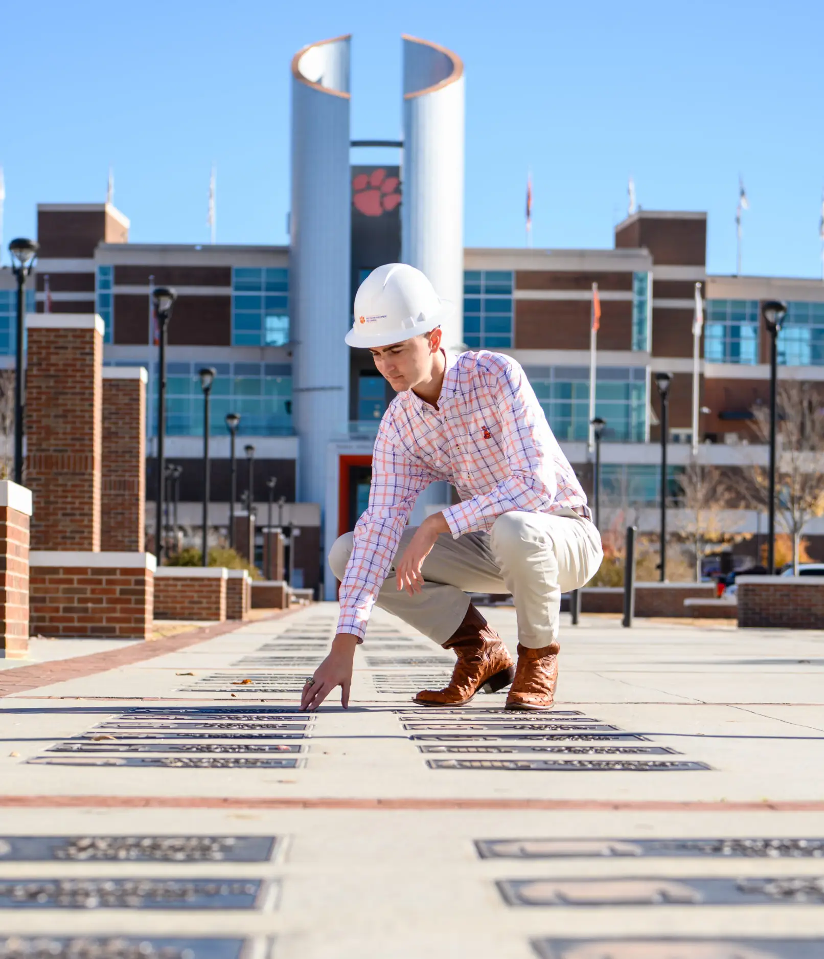 A male student wearing brown boots, khaki pants, an orange-and-purple checkered button-down shirt and a white construction hard hat kneels down in front of rows of inscribed plaques that are placed on a concrete walkway.