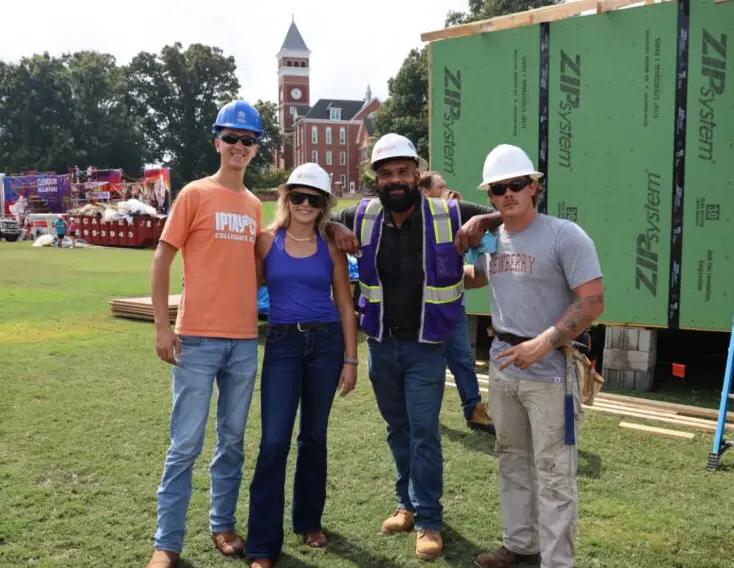 A group of two male students, one female student and a male professor, all wearing construction hard hats, stand on Bowman Field in front of the wooden frame of a house that is under construction. A pickup truck, construction materials, a brick clock tower and trees are visible in the background.