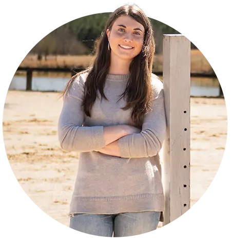 A female student wearing a gray sweater and denim jeans stands in an outdoor equestrian ring.