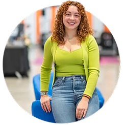 A female student with a lime green tank top and matching cardigan sweater and denim jeans leans on the back of a chair.