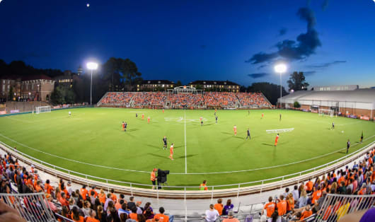 Athletes on a field and a bench with fans as a soccer game is being played.